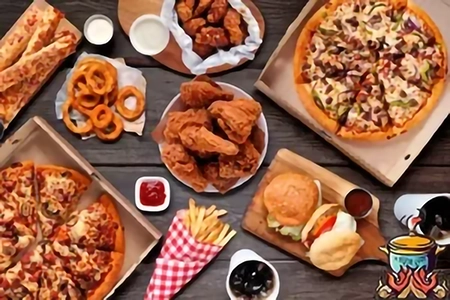 Vue de dessus d’une table en bois garnie de plats de restauration rapide : pizzas en parts, seaux de poulet frit croustillant, onion rings dorés, hamburgers avec garnitures, frites en cornet rouge et boisson gazeuse.