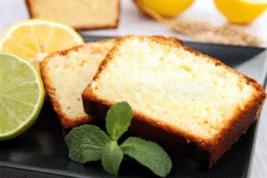 Tranches de cake au citron moelleux disposées sur une assiette noire, accompagnées de rondelles de citron vert et de citron jaune, avec une feuille de menthe en décoration.
