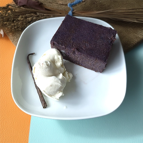 Assiette blanche contenant une part de gâteau de patate douce violette à la texture dense, accompagnée d’une boule de glace vanille et d’une gousse de vanille en décoration