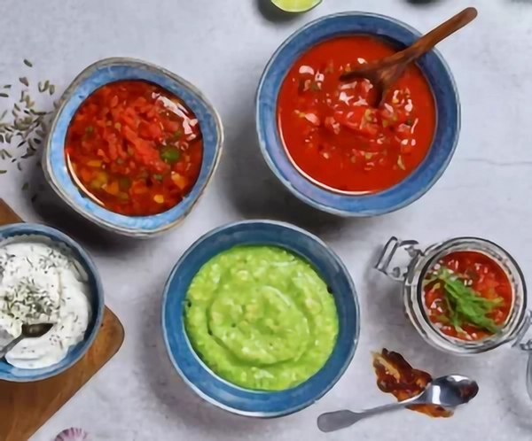 Assortiment de sauces maison dans des bols bleus et en verre : sauce tomate épaisse, sauce verte crémeuse type guacamole, sauce blanche au yaourt avec herbes, et une sauce tomate garnie de piments.