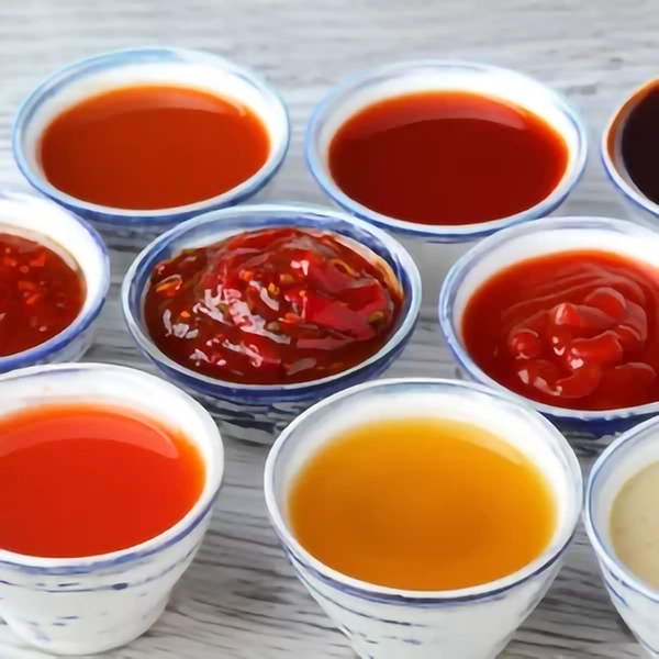 Assortiment de petits bols blancs alignés, contenant différentes sauces liquides et épaisses aux teintes rouges, orangées et brunes, comme ketchup, piment, sauce soja ou vinaigre.