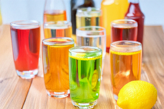 Plusieurs verres remplis de boissons fraîches sans alcool aux couleurs vives — vert, rouge, jaune, orange — posés sur une table en bois, décorés de rondelles de citron avec un citron entier en avant-plan.