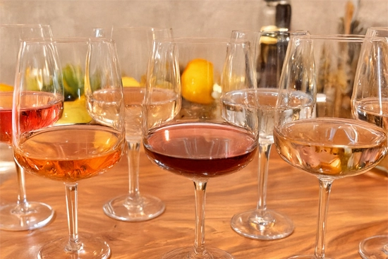 Assortiment de verres à vin posés sur une table en bois, remplis de différentes couleurs de vin : rouge profond, rosé clair et blanc transparent, avec en arrière-plan des citrons et bouteilles floutées.