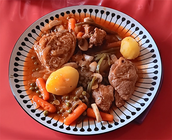 Assiette de sauté de veau aux petits légumes, servie dans une sauce tomate légère. On distingue des morceaux de viande, des pommes de terre entières, des carottes, des haricots verts, des champignons et des petits pois, le tout nappé d’une sauce rouge parfumée. L’assiette blanche est décorée de motifs noirs en forme de gouttes.