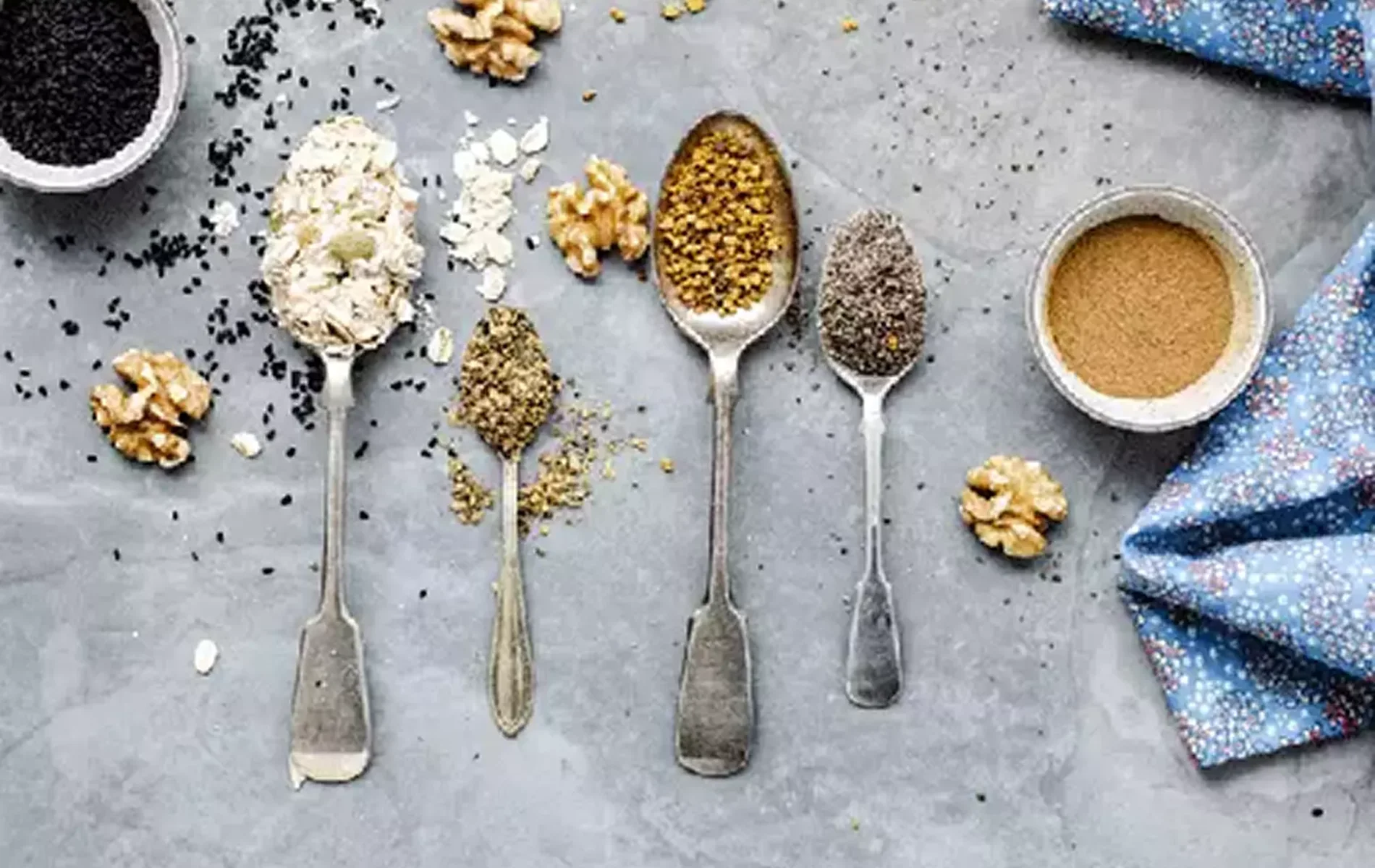 Quatre cuill&egrave;res anciennes align&eacute;es contenant flocons d&rsquo;avoine, &eacute;pices moulues, graines et noix concass&eacute;es, pos&eacute;es sur une surface grise avec quelques noix autour.