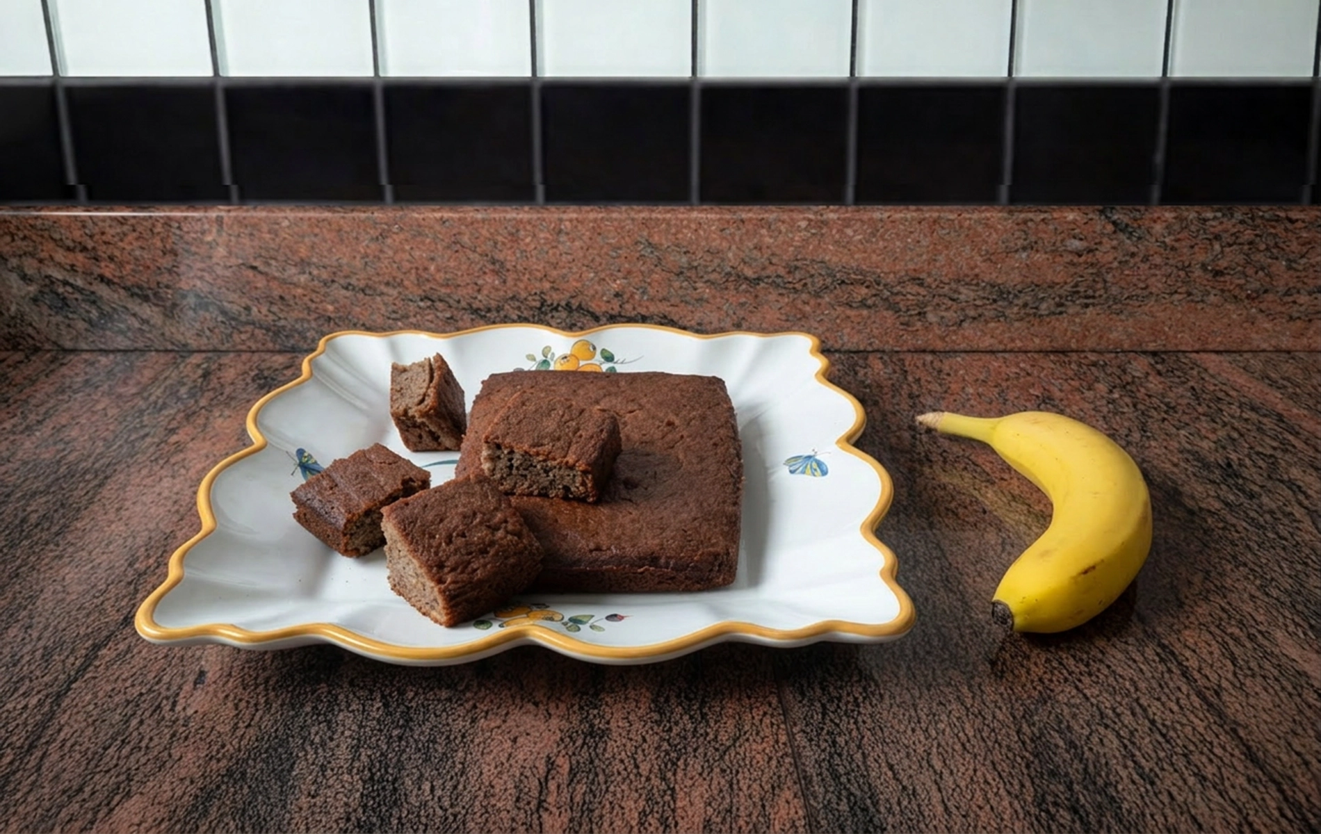G&acirc;teau moelleux &agrave; la banane coup&eacute; en plusieurs carr&eacute;s et pr&eacute;sent&eacute; sur un plat d&eacute;cor&eacute;, avec une banane enti&egrave;re &agrave; droite.