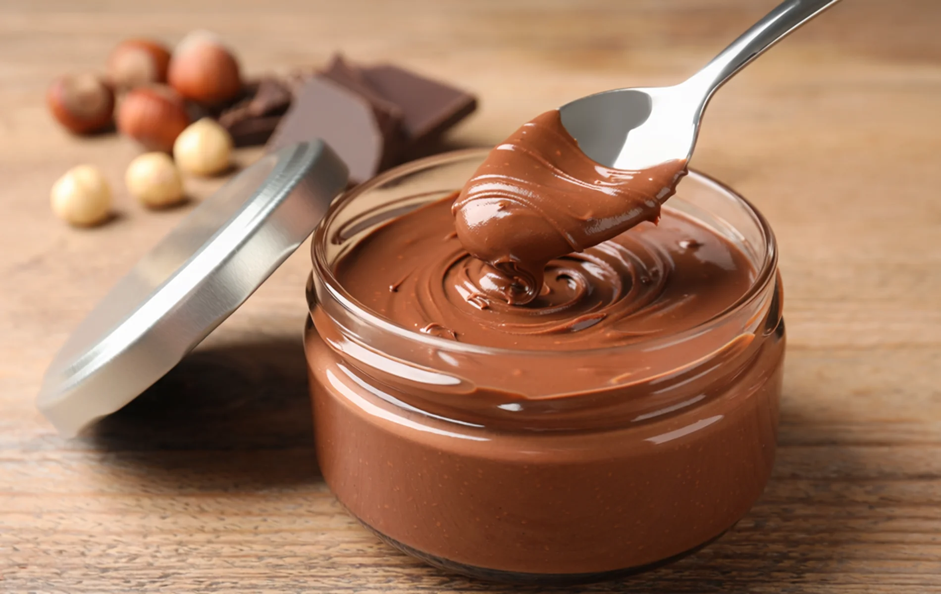 Un bocal ouvert de pâte à tartiner chocolat-noisette onctueuse, sur un fond de noisettes entières et carrés de chocolat.