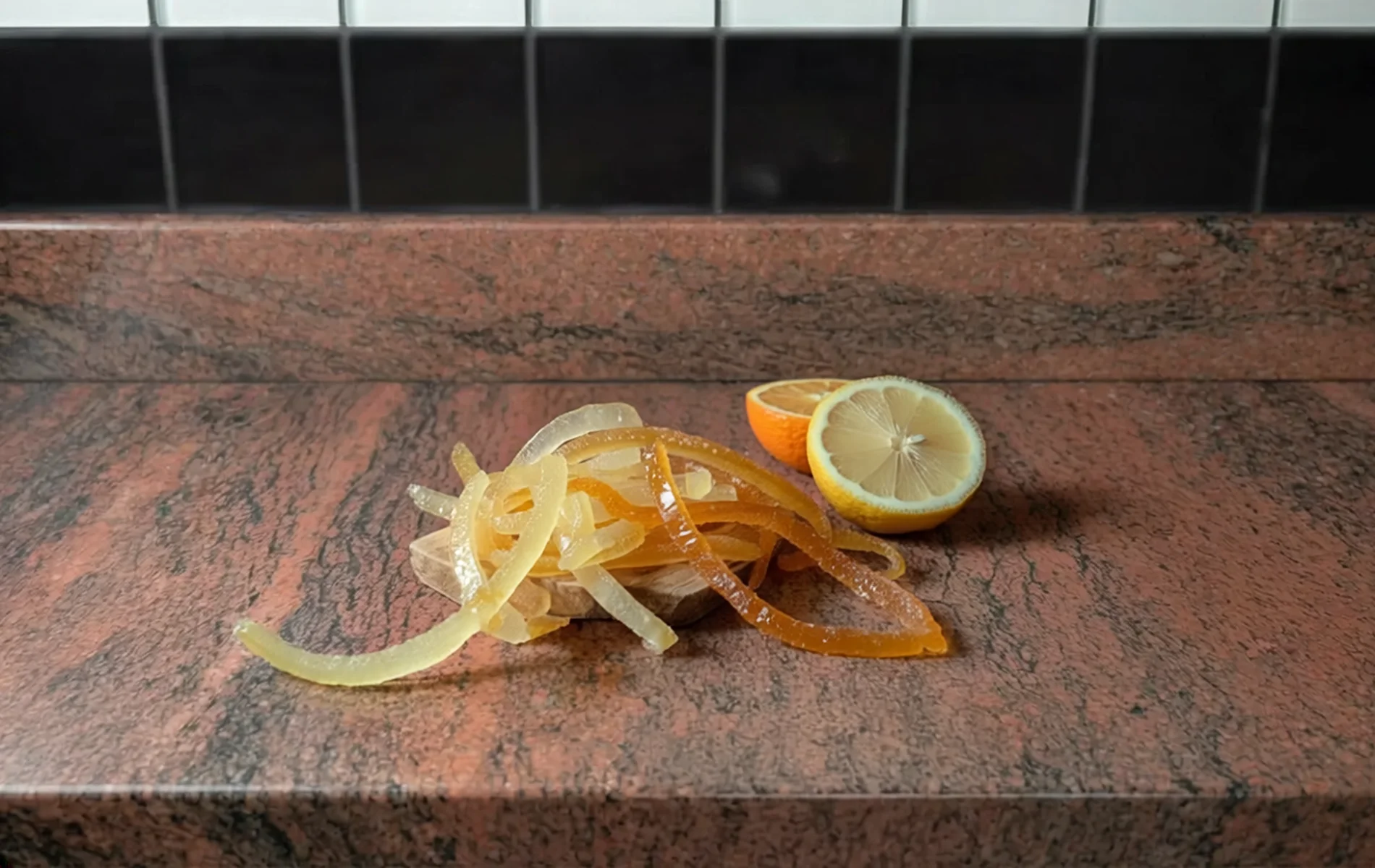 Zestes d’agrumes confits (citron et orange) en lamelles brillantes et translucides, posés sur une surface en pierre.