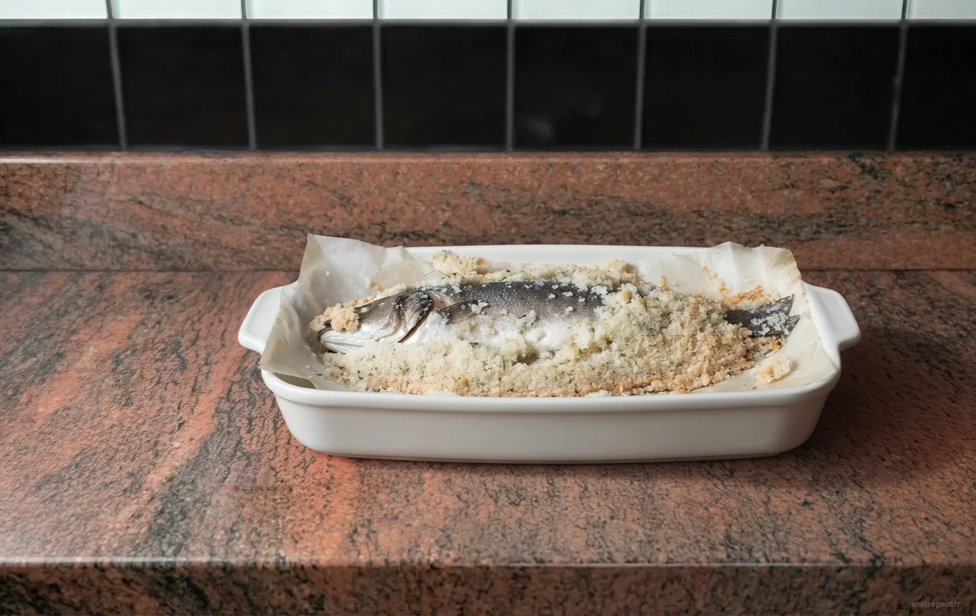 Bar en croûte de sel légèrement ouverte dans un plat ovale en céramique blanche, posé sur un plan de travail en granite
