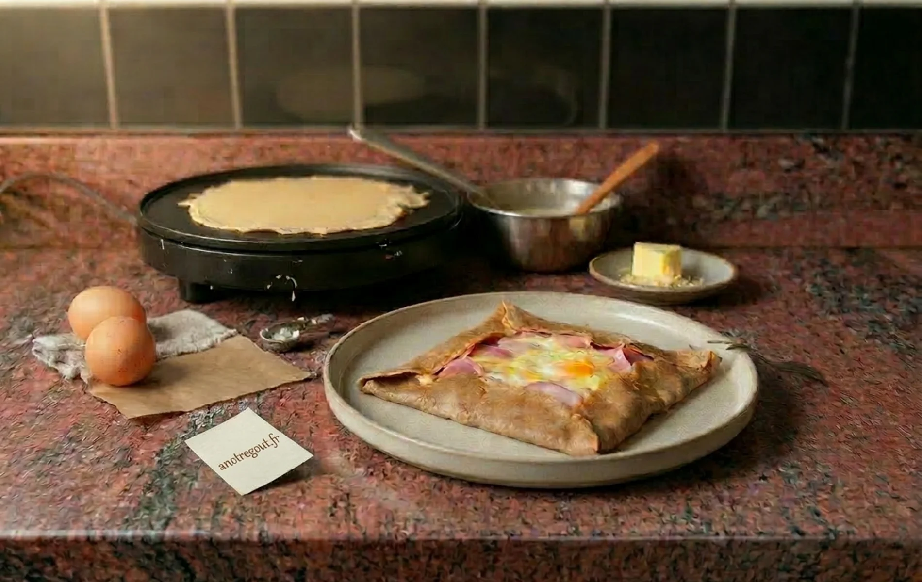 Galette de sarrasin garnie (œuf, jambon, fromage) dans une assiette, avec crêpière en arrière-plan.
