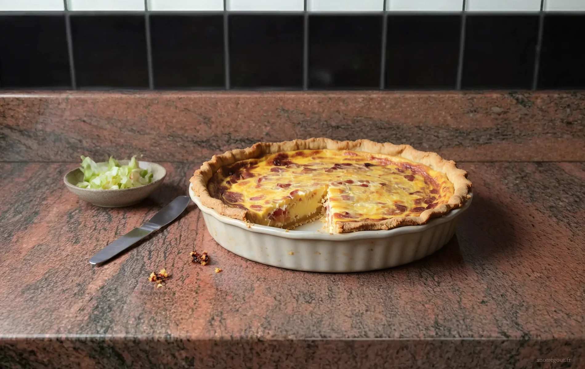 Quiche Lorraine avec part découpée, salade verte et couteau sur plan de travail en granit.