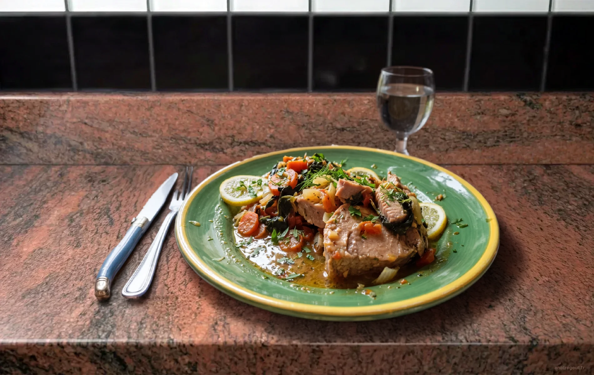 Thon en chartreuse aux l&eacute;gumes confits, citron et fenouil, servi dans une assiette verte avec couverts et verre en arri&egrave;re-plan