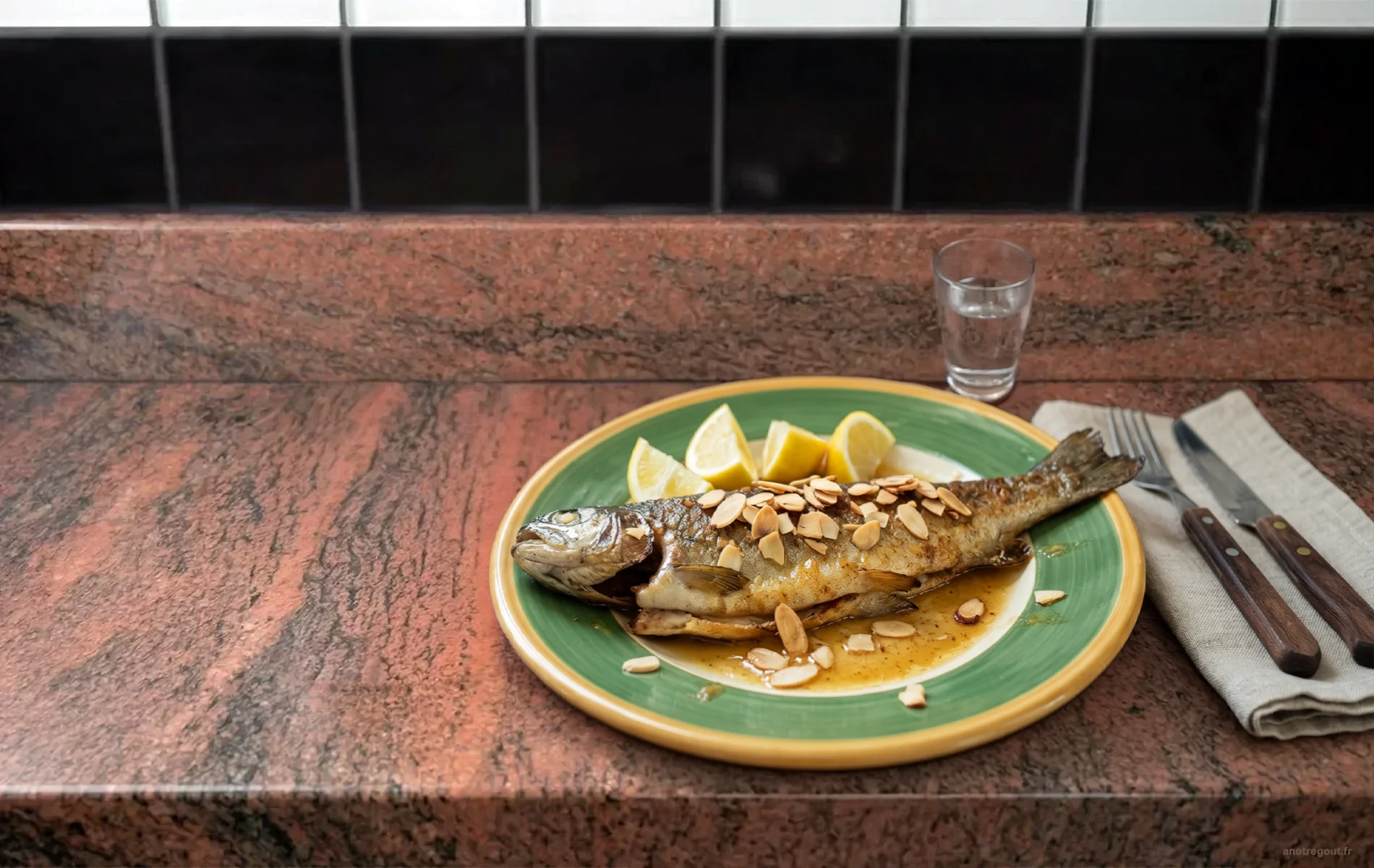 Truite aux amandes enti&egrave;re, beurre citronn&eacute; et quartiers de citron, servie dans une assiette verte avec couverts et verre en arri&egrave;re-plan