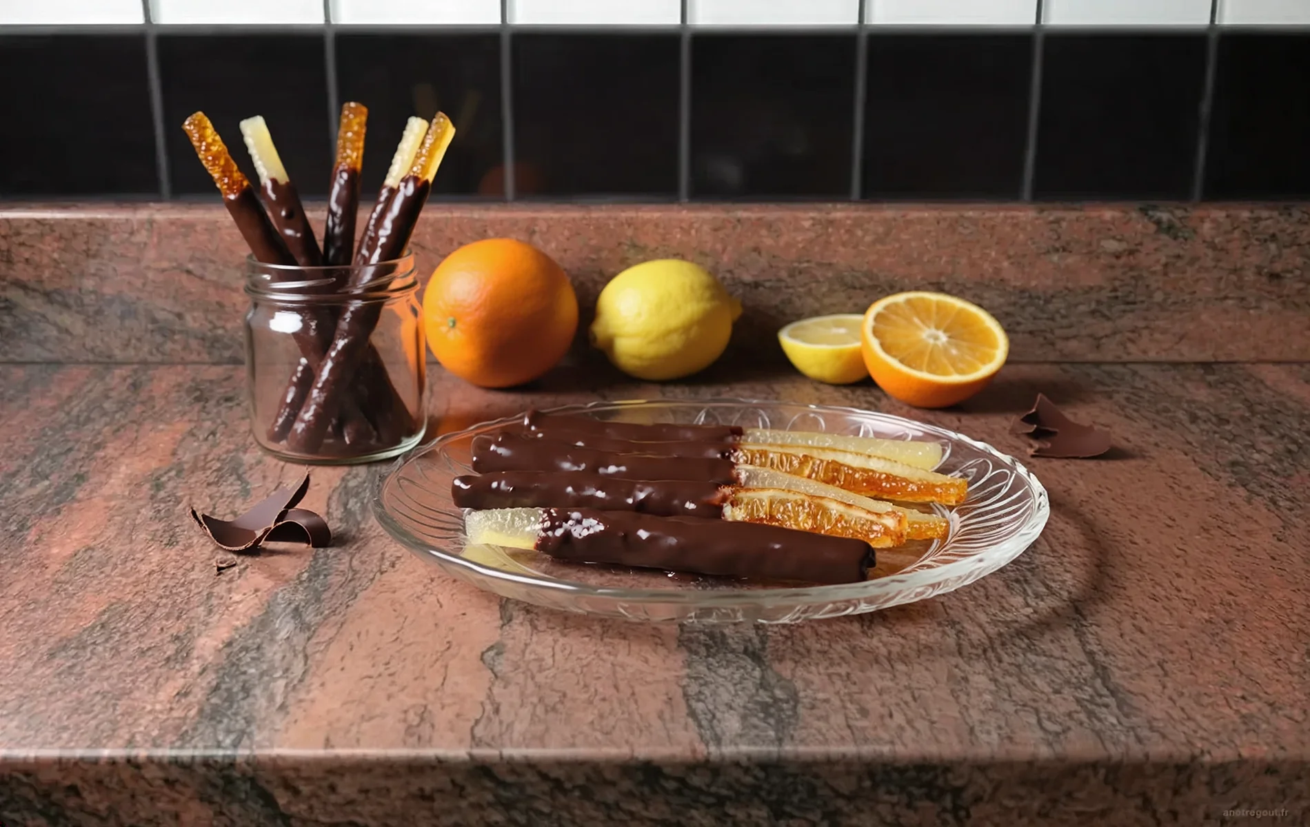 &Eacute;corces d'orange et de citron confites partiellement enrob&eacute;es de chocolat noir, pr&eacute;sent&eacute;es sur une assiette avec un bocal et des agrumes frais en arri&egrave;re-plan.