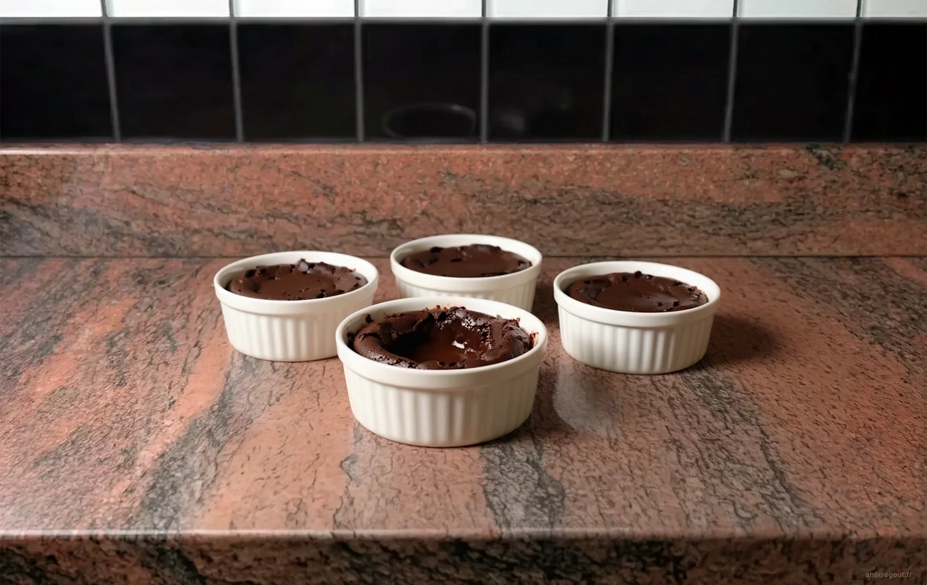 Quatre ramequins blancs de fondants au chocolat, dont un ouvert au premier plan révèle un cœur chocolaté brillant, sur un plan de travail en granit rose veiné.