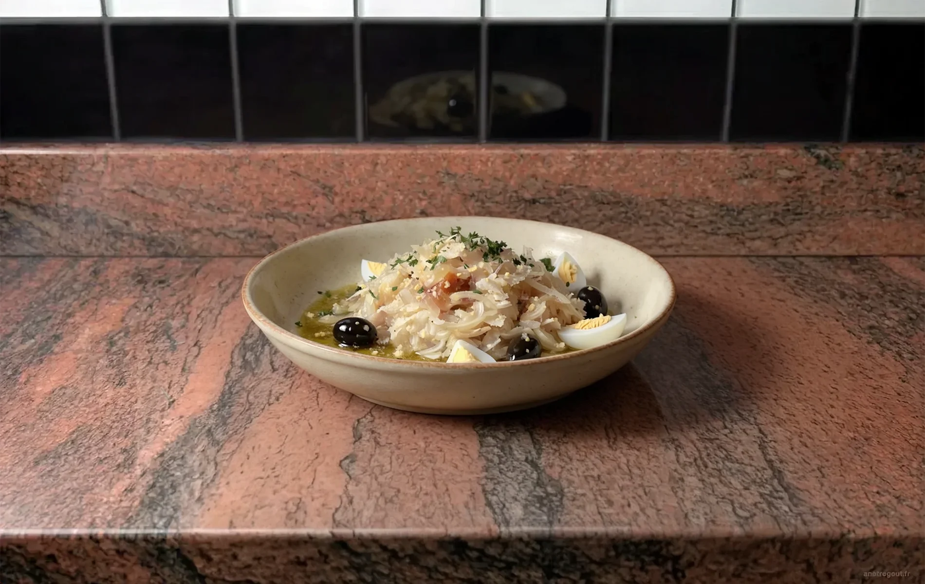 Salade portugaise de morue dessalée et effilochée, accompagnée d'olives noires, de quartiers d'œufs durs, d'oignons et de persil, dans un plat en céramique.